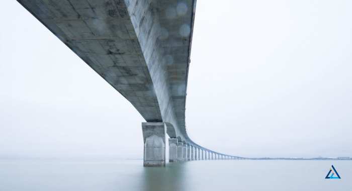 The image shows a bridge over a body of water. The structure is made of concrete and extends into the horizon. The main view of the bridge is its underside, displaying the concrete support pilars and the underside of the road above.